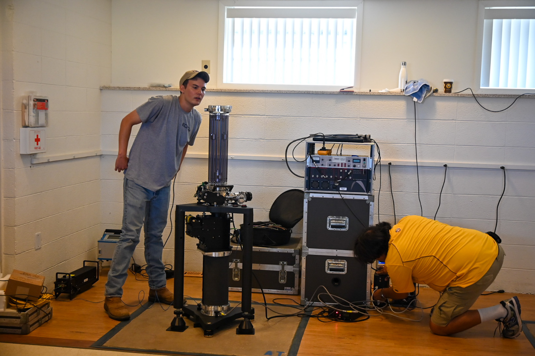 One man looks at the top of the gravity meter, inspecting a level, while another kneels beside it plugging in various cords.