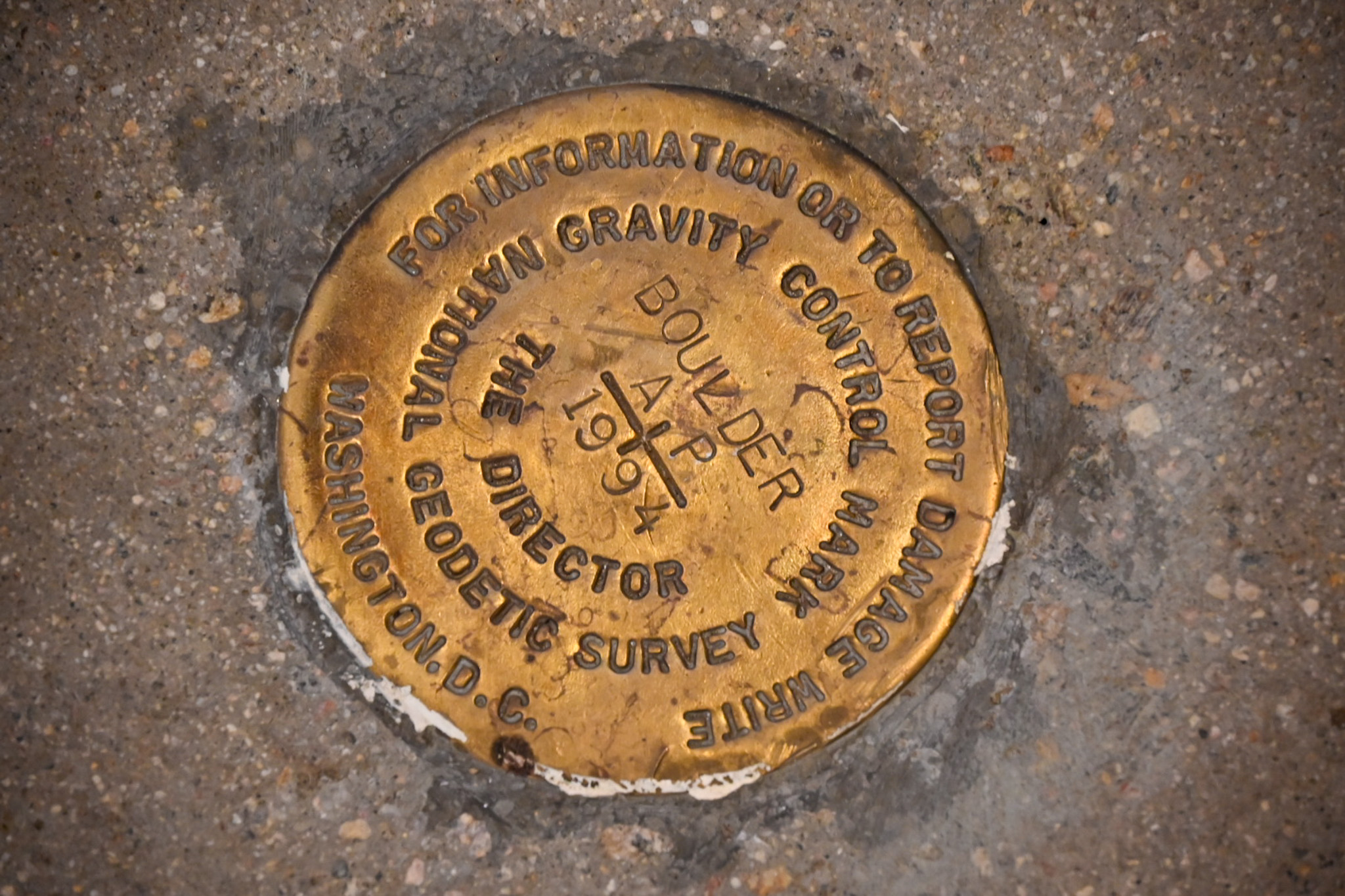 A metal round marker is embedded into cement. The marker reads, “ Gravity Control Mark,For information or to report damage write the director, National Geodetic Survey, Washington, D.C.”
